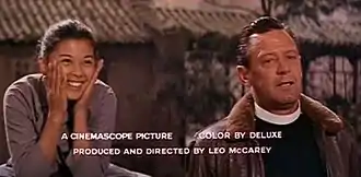 An East Asian young woman smiling with her hands on her cheeks and a white priest in a pilot jacket. A wooden building is in the background. In the foreground, the text "A CINEMASCOPE PICTURE  COLOR BY DELUXE / PRODUCED AND DIRECTED BY LEO MCCAREY