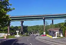 A pair of bridges pass from left to right on tall supports that tower over the other structures in the area, such as telephone poles and single-story buildings. The two-lane NY&nbsp;22 passes underneath the bridge and proceeds into the background.