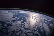 Image 32A view of Earth with different layers of its atmosphere visible: the troposphere with its clouds casting shadows, a band of stratospheric blue sky at the horizon, and a line of green airglow of the lower thermosphere around an altitude of 100 km, at the edge of space (from Earth)