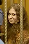 A woman with strawberry blonde curly long hair smile toward the camera. The photo is obstructed by two bars of a very small cell.