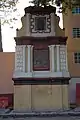 Picture of a three-story monument. From bottom to top, the foundation is a relieved wall; the next story features a depiction of the announcement by the Archangel Gabriel to the Virgin Mary that she would conceive and become the mother of Jesus. The last story features an image of Our Lady of Guadalupe.