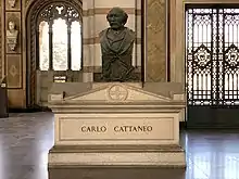 A white marble sarcophagus, surmounted by a bronze bust of the deceased, inside a chapel