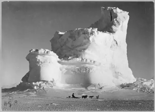 Photo of a three-tiered iceberg resembling a fanciful castle, dwarfing a sled team in the foreground